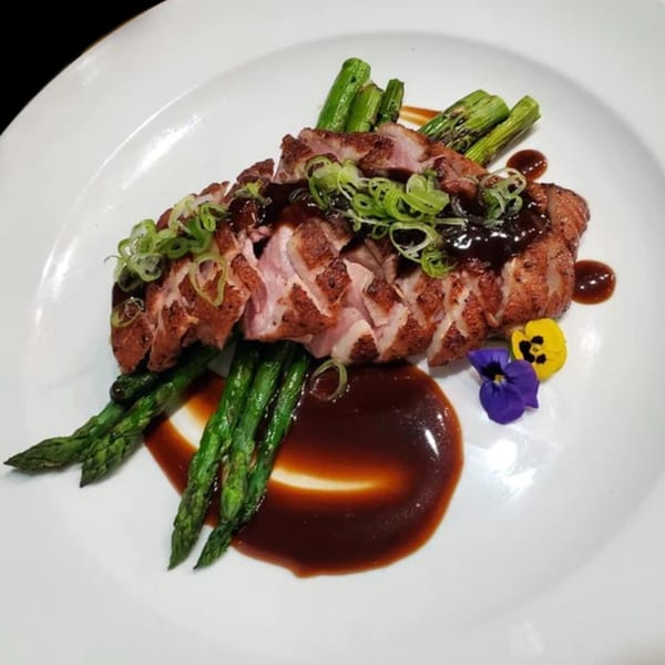 Sliced duck breast with dark sauce, green onions, asparagus, and edible flowers on a white plate.