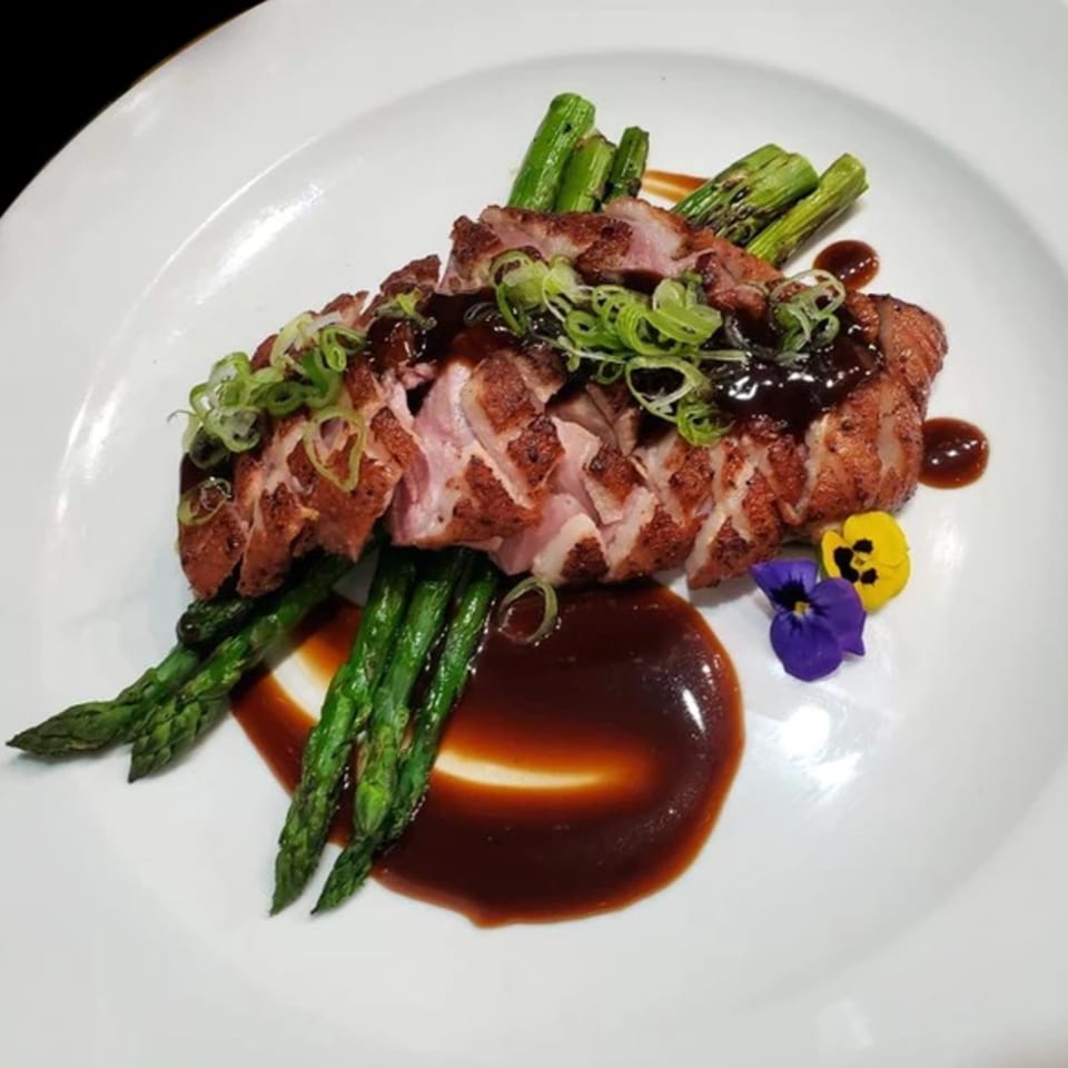 Sliced duck breast with dark sauce, green onions, asparagus, and edible flowers on a white plate.