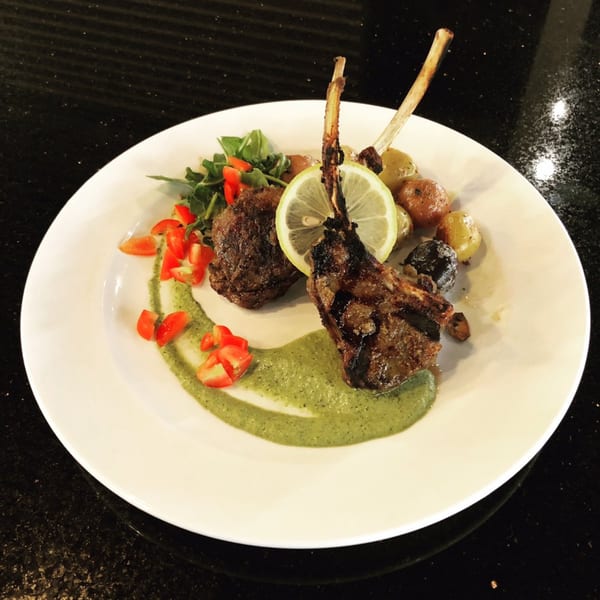 Grilled lamb chops with lemon, roasted potatoes, greens, and chopped tomatoes on a white plate.